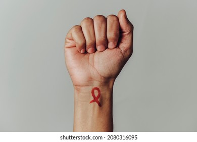 the raised fist of a man with a red awareness ribbon for the fight against AIDS painted in his wrist, against an off-white background with some blank space around him