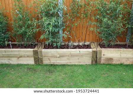 Raised flower or vegetable beds, or planters, or troughs made with wooden railway sleepers. Green lawn, orange fence and bamboo screening. Outdoors on a spring day