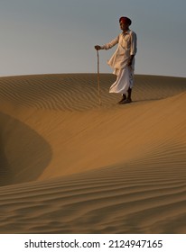 Rajasthan India February 2020: A Nomadic Camel Herder Goes For A Walk As The Sun Sets In The Thar Desert 