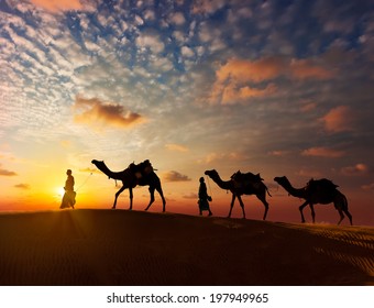 Rajasthan travel background - two indian cameleers (camel drivers) with camels silhouettes in dunes of Thar desert on sunset. Jaisalmer, Rajasthan, India - Powered by Shutterstock