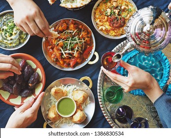 Ramadan Iftar Meal Theme People Eating Variety Foods Together On Dining Table With Large Dates (Medjool), Samosas, Aloo Gobi, Chicken Biryani, Moroccan Tea Set, Palak Paneer And Naan Bread. (top View)