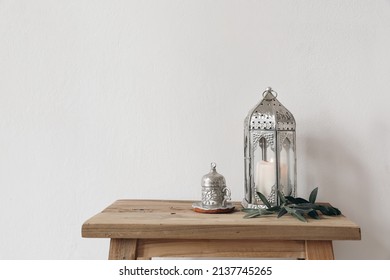 Ramadan Kareem still life. Ornamental lantern with burning candle and Turkish silver cup with tea or coffee. Green olive tree branches on old wooden table background. Muslim Iftar dinner. No people
