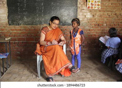 Ramgarh, Bihar, India - April 23 2019: Indian Teacher Woman In Orange Saree Sitting On A Chair In A Rural Village School And Checking A Workbook, Little Girl Student Stands Near