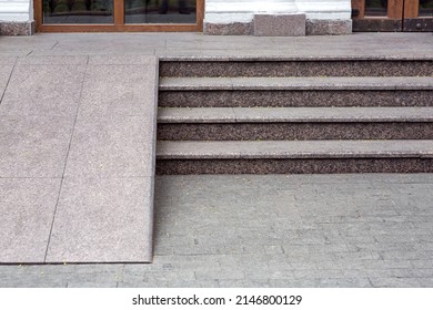 Ramped access, using wheelchair ramp for disabled people. Granite ramp pathway near stone tile stairs of entrance to cafe with glass door close-up front view, nobody.