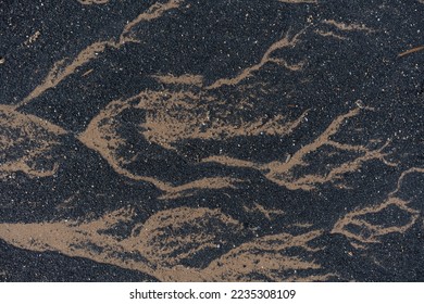Random natural patterns made by water retreating across dark gritty sand in a river estuary. The texture is black and tan with the detail running horizontally. Small stones and grit dot the surface.