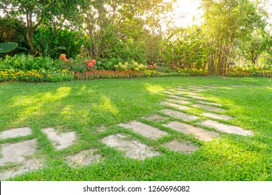 Random Pattern Of Grey Concrete Stepping Stone On Green Grass Lawn, Flowering Plant, Shrub And Trees On Backyard Under Morning Sunshine With Good Care Landscaping In A Garden Of The Public Park  