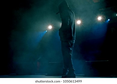 Rap Artist On Stage In The Rays Of Soffits Light. Concert Backlight And Illumination During Music Concert. Singer In Hoodie With Microphone At Stage