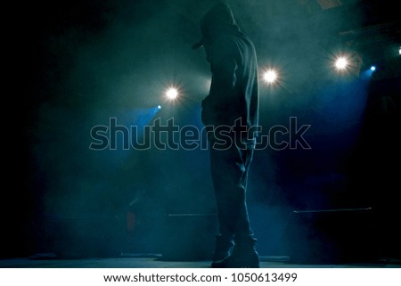 Rap artist on stage in the rays of soffits light. Concert backlight and illumination during music concert. Singer in a hoodie with a microphone on the stage. Musicant start a concert 