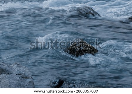Rapid river currents creating a small white wave and vortex around a dark stone in a close-up shot of flowing water.