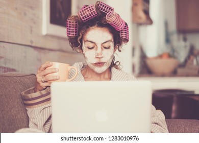 Rare and funny portrait of beautiful woman with facial mask and curlers hair taking a coffee at home while use internet with modern laptop computer - home scene for modern lifestyle people 