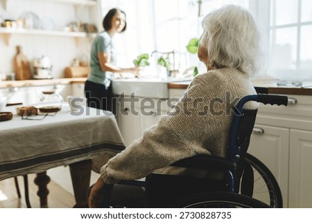 Rare view image with selective focus on old disabled female in wheelchair talking to her home assistant or caregiver cleaning kitchen or cooking dinner standing next to window in front of counter