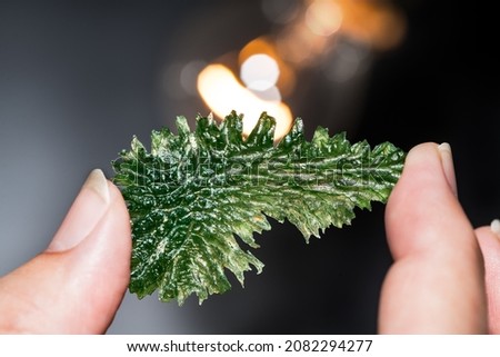 Rare wrinkled green moldavite mineral in human hand with orange light on black background. Collector holding meteoric glass gem from Czechia at incandescent lamp with beautiful bokeh. Selective focus.