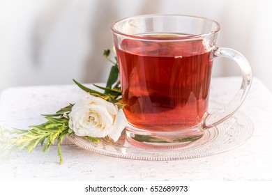 Rasberry Flavored Herbal Tea Served In A Glass Cup And Saucer With A White Rose On A White Rustic Wooden Table