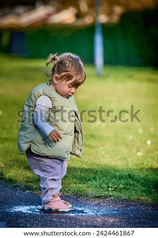 Rascal little female child with braids jumping in a muddy puddle on a sunny spring day. Scoundrel 2 year old girl hopping in a small water plash by the grass. Kids exploration and freedom concept
