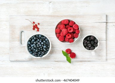 Raspberries, blackberries and bilberries in bowls served with fresh mint and red currant on white painted wooden background, top view, horizontal
