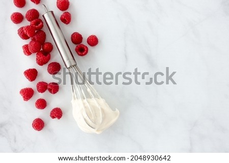Raspberries and a whisk with whipped cream on white marble background and lots of copy space. Top view.
