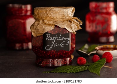 Raspberry Jam In Glass Jars, Ripe Raspberries On Dark Background. Organic Jam. Handwritten Label. Preserved Berries, Homemade Jam. 