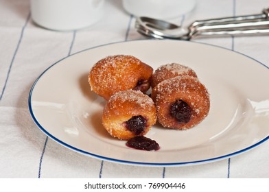 Raspberry Jelly Filled Brioche Donut Holes On A Round, White Plate With A Blue Rim On A White And Blue Tabel Cloth With Artisan Spoons And Two Coffee Cups