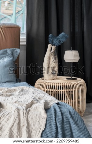 Rattan bedside table with decorative vase with blue feather and modern lamp next to cozy bed with blue and beige bedclothes
