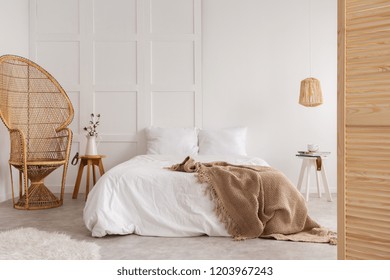 Rattan Chair And Wooden Table Next To Bed With Brown Blanket In White Bedroom Interior. Real Photo