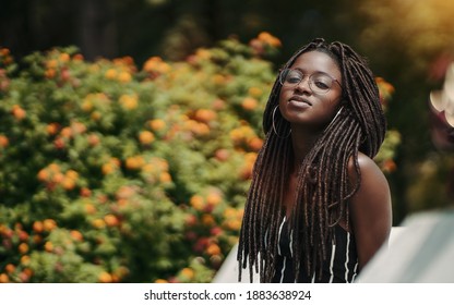 A ravishing young black female with dreadlocks and eyeglasses in the public park with a bush with flowers in the defocused background; a portrait of a dazzling African woman with dreads and in glasses