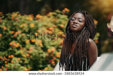 A ravishing young black female with dreadlocks and eyeglasses in the public park with a bush with flowers in the defocused background; a portrait of a dazzling African woman with dreads and in glasses