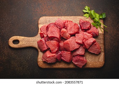 Raw Beef Meat Chopped In Cubes With Bunch Of Fresh Parsley On Wooden Cutting Board For Cooking Stew Or Other Meat Dish On Brown Dark Stone Concrete Background Top View Flat Lay