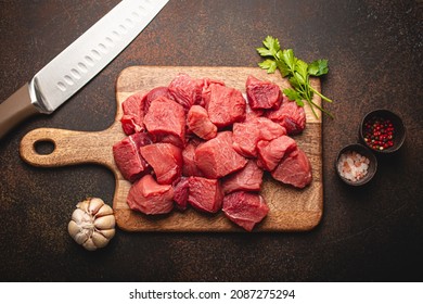 Raw beef meat chopped in cubes with bunch of fresh parsley, garlic, salt and pepper on wooden cutting board with knife for cooking stew or meat dish on brown dark stone background top view flat lay - Powered by Shutterstock