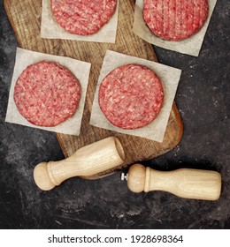 Raw Beef Pork Burgers, Grill Tools And Spice Mill On Wooden Board, Top View. Raw Farmers Ground Beef Meat Steak Burger Cutlets On Black Table Background, Overhead View. Homemade Uncooked Beef Burgers.