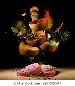 Raw Beef Steaks On Wooden Table With Various Spices And Herbs Flying With Splash Over Black Background