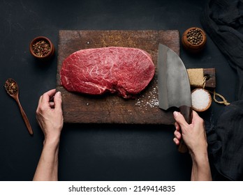 Raw beef tenderloin lies on a wooden cutting board and spices for cooking on a black table, top view. A woman's hand holds a large kitchen knife in her hand - Powered by Shutterstock