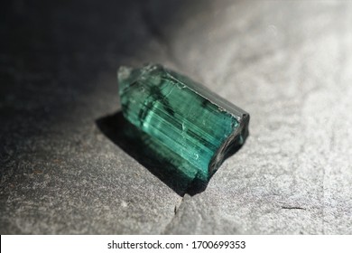 Raw blue-green tourmaline crystal, on gray slate background, lit by the sun