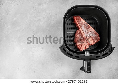Raw bone in beef steak placed in an air fryer basket on a light concrete surface, illustrating modern home cooking and meal preparation.