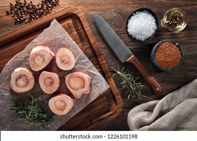 Raw calf bones on white cooking paper and wooden cutting board. Decorated with herbs, spices, chef's knife and napkin. Overhead view.
