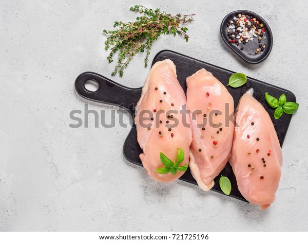 Raw chicken breast with fresh basil and\
thyme on black cuttingboard. Raw chicken fillet on gray cement\
background. Copy space. Top view or flat\
lay.