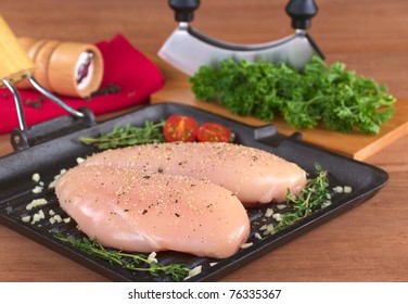 Raw Chicken Breast In Frying Pan Seasoned With Pepper, Garlic And Thyme With Fresh Herb And Mezzaluna As Well As A Pepper Mill In The Back (Selective Focus, Focus On The Front Of The Meat)