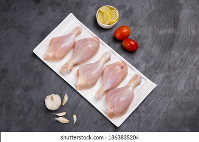 Raw chicken drum sticks arranged on white table ware with tomato ,lemon slices and garlic on grey colour background, top view