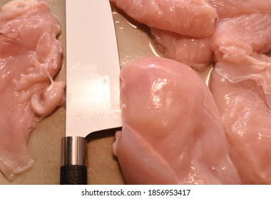 The Raw Chicken Fillet, From Which The Juice Drips, Lies On A Cutting Board. Nearby There Is A Large Ceramic Knife.