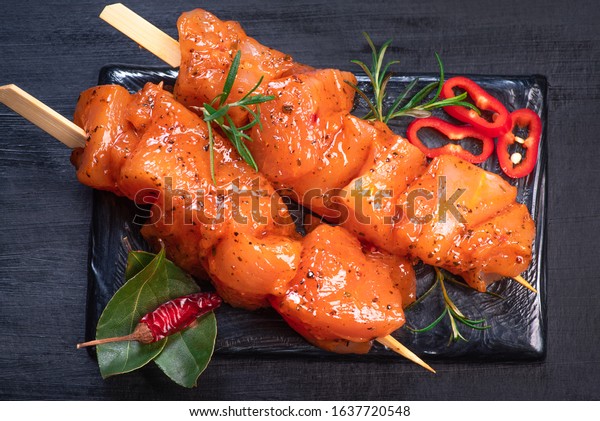 Raw chicken skewers in marinade with spices on a\
black plate and on a wooden table. Raw marinated and spicy chicken\
skewers.Top view. Chicken meat close up. Raw meat in marinade.\
Tasting diet meat.