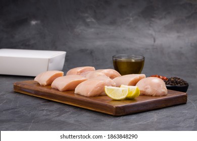 Raw Chicken Tender Fry Cut Without Skin Arranged On Wooden Board And Garnished With Oil,lemon Slices , Chilly Flakes And Black Pepper On Stone Textured Or Graphite Colour Background With Take Away Box