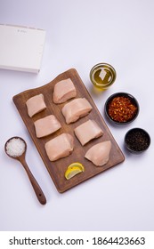 Raw Chicken Tender Fry Cut Without Skin Arranged On Wooden Board And Garnished With Oil,lemon Slices , Chilly Flakes And Black Pepper On White Colour Background With Take Away Box