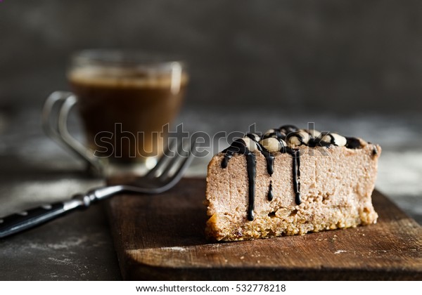 Raw chocolate mousse cake  with cashew nuts,\
hazelnuts and dark chocolate glaze topping and coffee on a wooden\
and grey stone background. Vegan sugar, gluten, dairy free dessert.\
Horizontal
