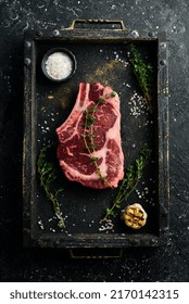 Raw Cowboy Steak With Spices On A Stone Background, Prime Ribeye On The Bone. Top View. On A Black Stone Plate.