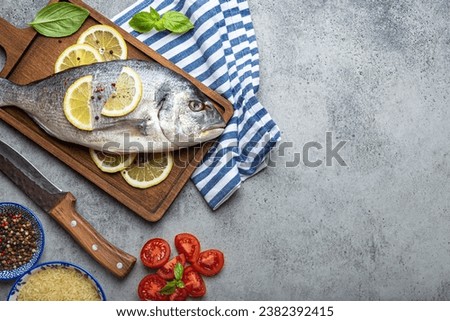 Raw fish dorado with ingredients lemon, fresh basil, cut cherry tomatoes, uncooked rice on wooden cutting board with knife on rustic stone table top view, cooking healthy fish dorado. Copy space
