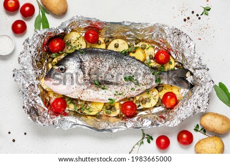 Raw fish with potatoes, tomatoes, and herbs in foil. Uncooked dorada (gilt-head bream) with vegetables in foil on a gray concrete background top view.