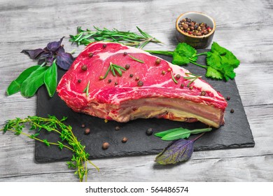 Raw fresh cow meat rib eye steak on the black stone board. Seasoning for meat rib eye steak thyme, basil, sage, rosemary, coriander, pepper. Light white wooden background