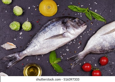 Raw Fresh Dorado Fish With Brussels Sprouts, Tomatoes, Lemon, Young Potato, Greens, Bread, White Wine Bottle And Olive Oil On Dark Background. View From Above, Top Studio Shot