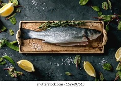 Raw Fresh Uncooked Fish Sea Bass On Ceramit Plate Ready To Cook With Lemon, Sea Salt, Rosemary, Green Salad Over Dark Texture Concrete Background. Flat Lay, Space