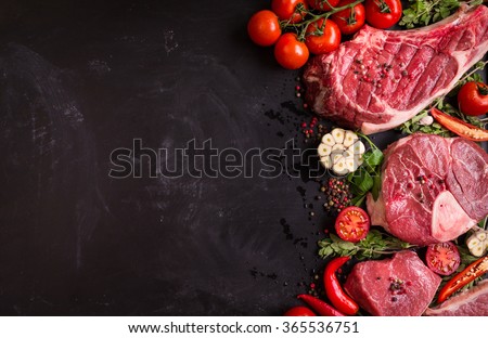Raw juicy meat steaks ready for roasting on a black chalk board background. Rib eye steak on the bone, veal shank (ossobuco), fillet with cherry tomatoes, hot pepper and herbs. Space for text