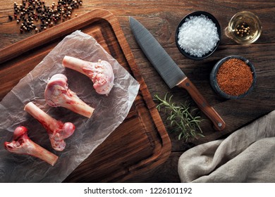 Raw Lamb Bones On White Cooking Paper And Wooden Cutting Board. Decorated With Herbs, Spices, Chef's Knife And Napkin. Overhead View.